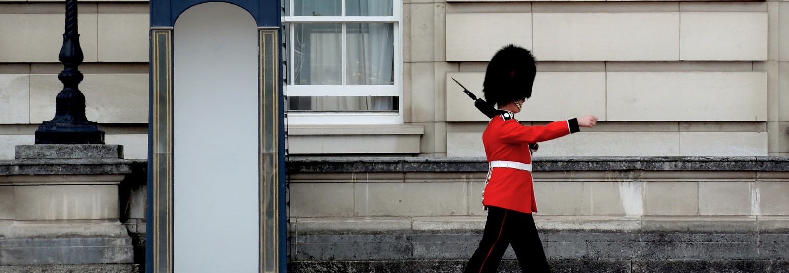 British guard in red jacket and tall black hat marching in front of a building.
